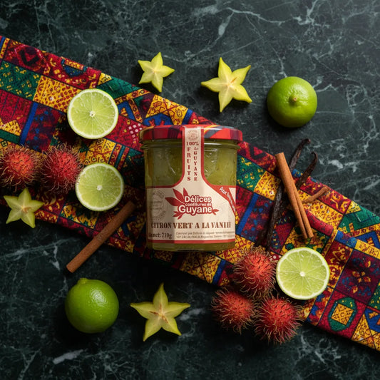 Pot de confiture Citron Vert Vanille DÉLICES DE GUYANE entouré de fruits exotiques comme ramboutans, citrons verts et caramboles sur tissu coloré.