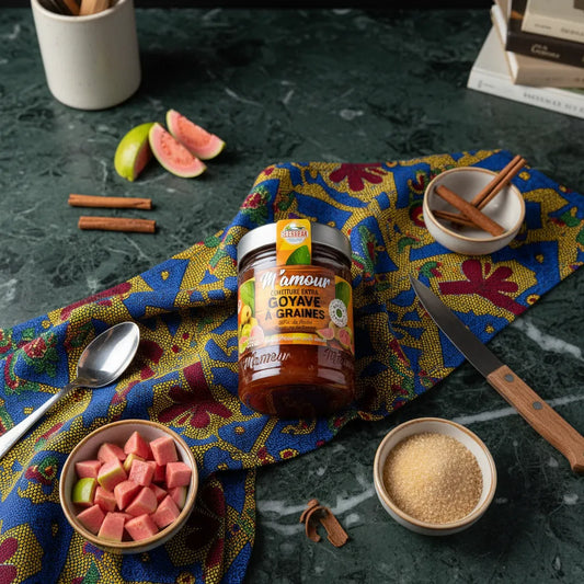 Pot de confiture de goyave M'AMOUR posé sur un tissu coloré avec morceaux de goyave, sucre roux et cannelle, ambiance épicerie sucrée authentique.