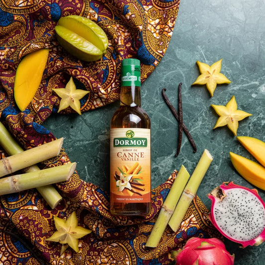 Sirop vanille canne DORMOY entouré de fruits tropicaux et gousses de vanille, arôme unique pour condiments et sauces sucrées.