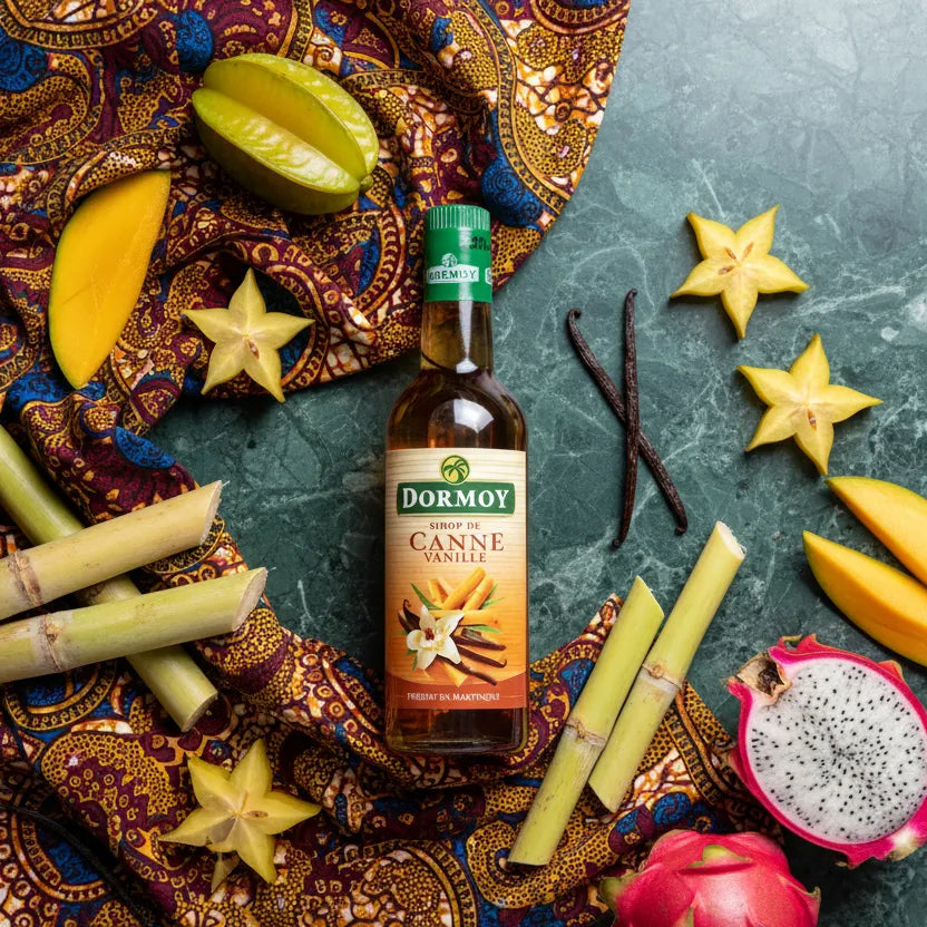 Sirop vanille canne DORMOY entouré de fruits tropicaux et gousses de vanille, arôme unique pour condiments et sauces sucrées.