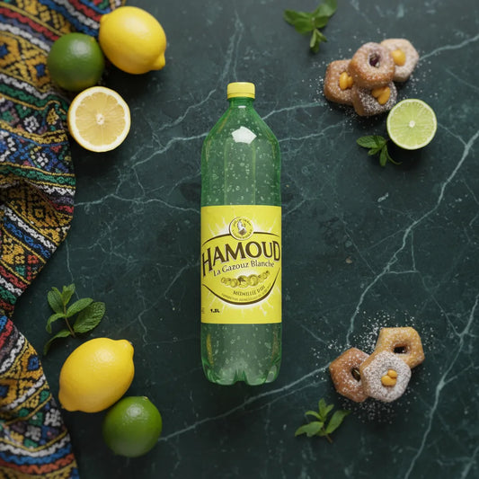 Bouteille de limonade HAMOUD entourée de citrons, limes et pâtisseries, boisson gazeuse idéale pour accompagner les douceurs orientales.