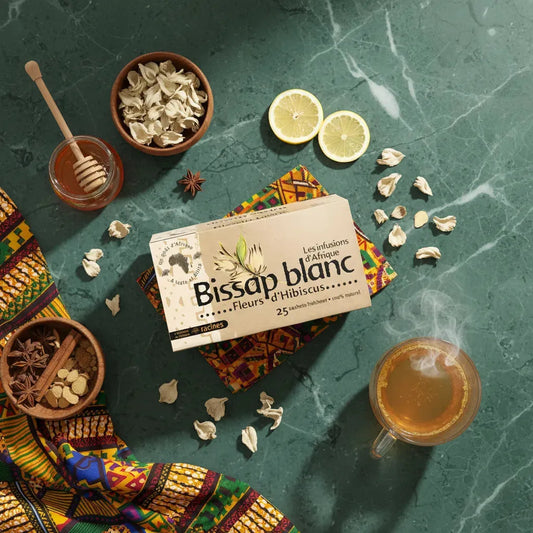 Infusion bissap blanc RACINES présentée avec citron, miel et fleurs séchées sur fond vert et tissu africain, idéale chaude ou froide.