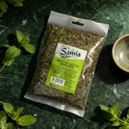 Sachet de menthe douce brisure SAMIA posé sur un plan de travail vert, herbes fraîches visibles, parfait pour cuisine et infusions.