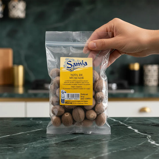 Main tenant un sachet de noix de muscade SAMIA sur un plan de travail de cuisine, épice entière d’Indonésie pour plats raffinés et épicés.