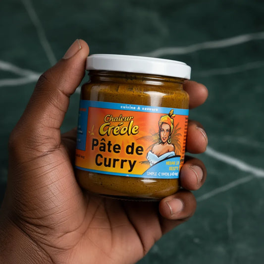 Main tenant un pot de Pâte de Curry Chaleur Créole, condiment épicé pour plats antillais. Étiquette vive et attrayante.