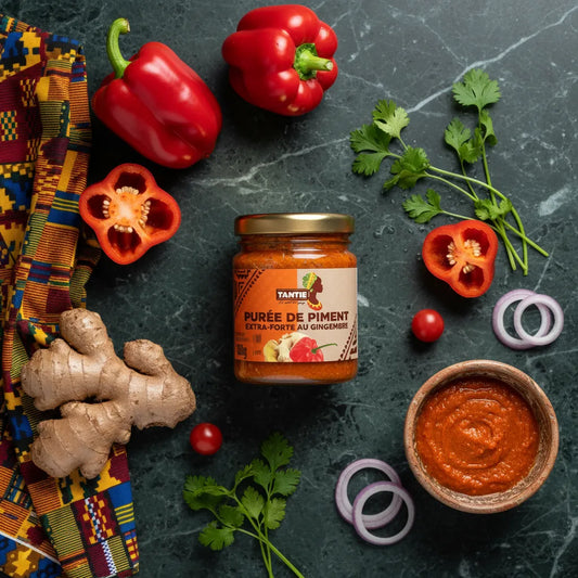 Pot de purée de piments gingembre TANTIE entouré d'ingrédients frais comme gingembre, poivrons et coriandre sur fond marbré.