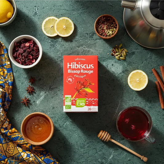 Boîte d'infusion Hibiscus Bissap Rouge BIO RACINES sur table avec ingrédients naturels, citron, miel et épices pour une boisson bio.
