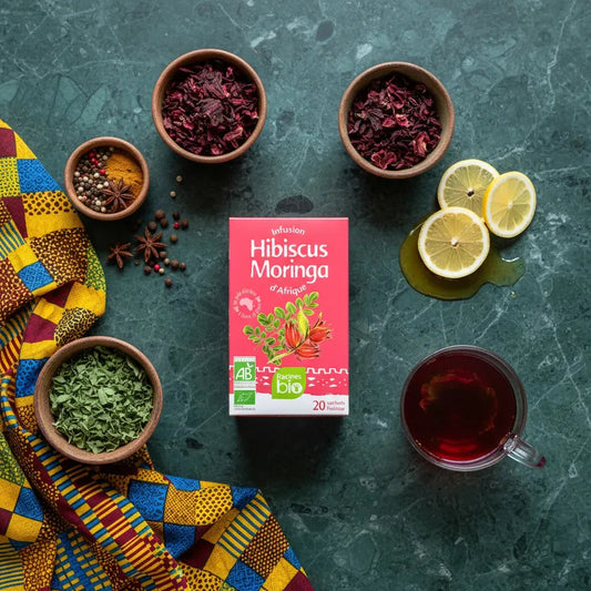 Infusion hibiscus moringa bio RACINES entourée d'ingrédients naturels : hibiscus séché, moringa, citron et épices sur fond vert marbré.
