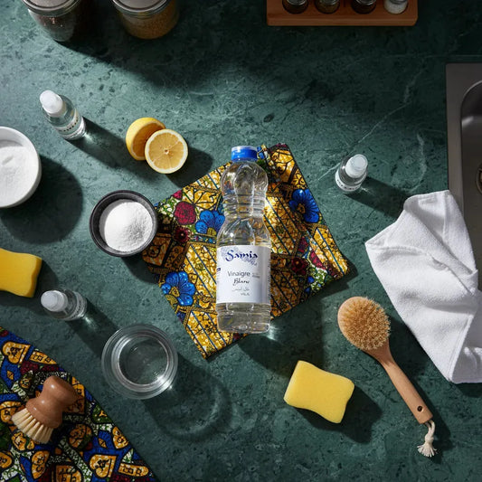 Bouteille de vinaigre blanc halal SAMIA posée sur un tissu coloré, entourée d’ustensiles de nettoyage et d’ingrédients naturels.