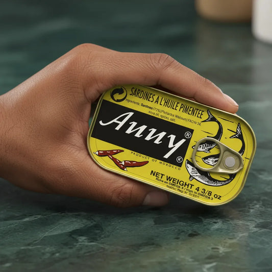 Main tenant une boîte de sardines épicées ANNY, mettant en avant le packaging jaune et noir au design attractif pour l'épicerie salée.