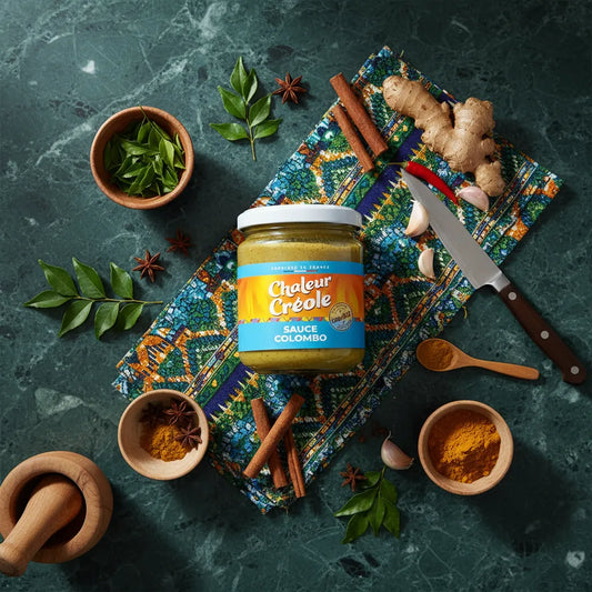 Pot de sauce colombo CHALEUR CREOLE posé sur un tissu créole avec épices fraîches comme curcuma, gingembre et cannelle autour.