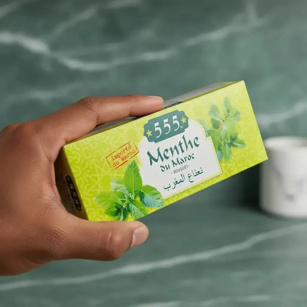 Main tenant une boîte de thé menthe sèche NANA 555, produit importé du Maroc, parfait pour une infusion digestive et rafraîchissante.