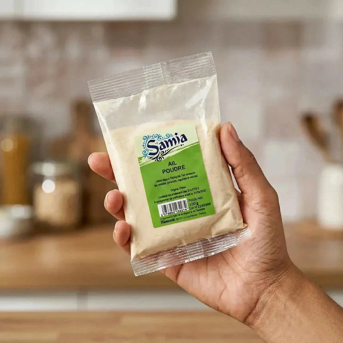 Un sachet de poudre d'ail SAMIA tenu à la main, fond neutre de cuisine. Étiquette verte et blanche.