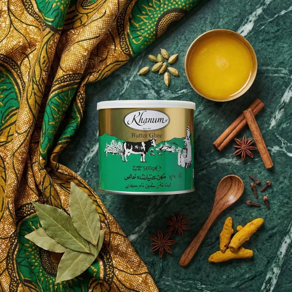 Khanum ghee butter in a tin (500g) surrounded by spices and colorful fabric on a green marbled background.