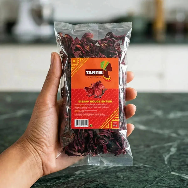 Sachet transparent Bissap rouge entier TANTIE tenu à la main sur fond vert marbré.