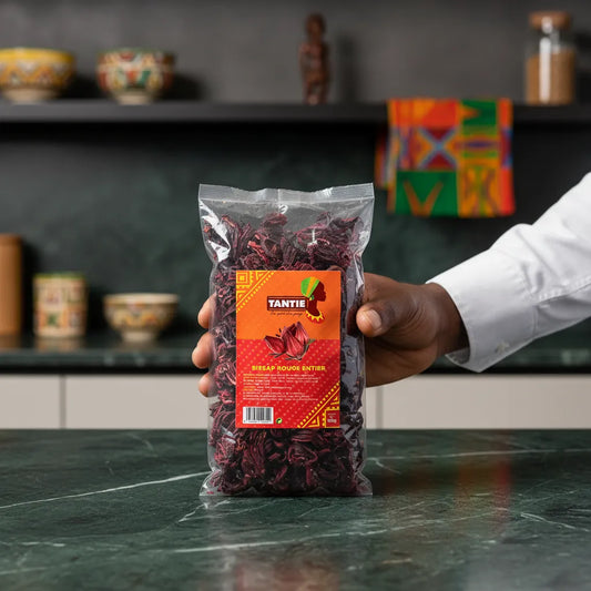 Main tenant un sachet de Bissap rouge TANTIE sur fond de cuisine africaine. Produit idéal pour infusions artisanales et boissons traditionnelles.