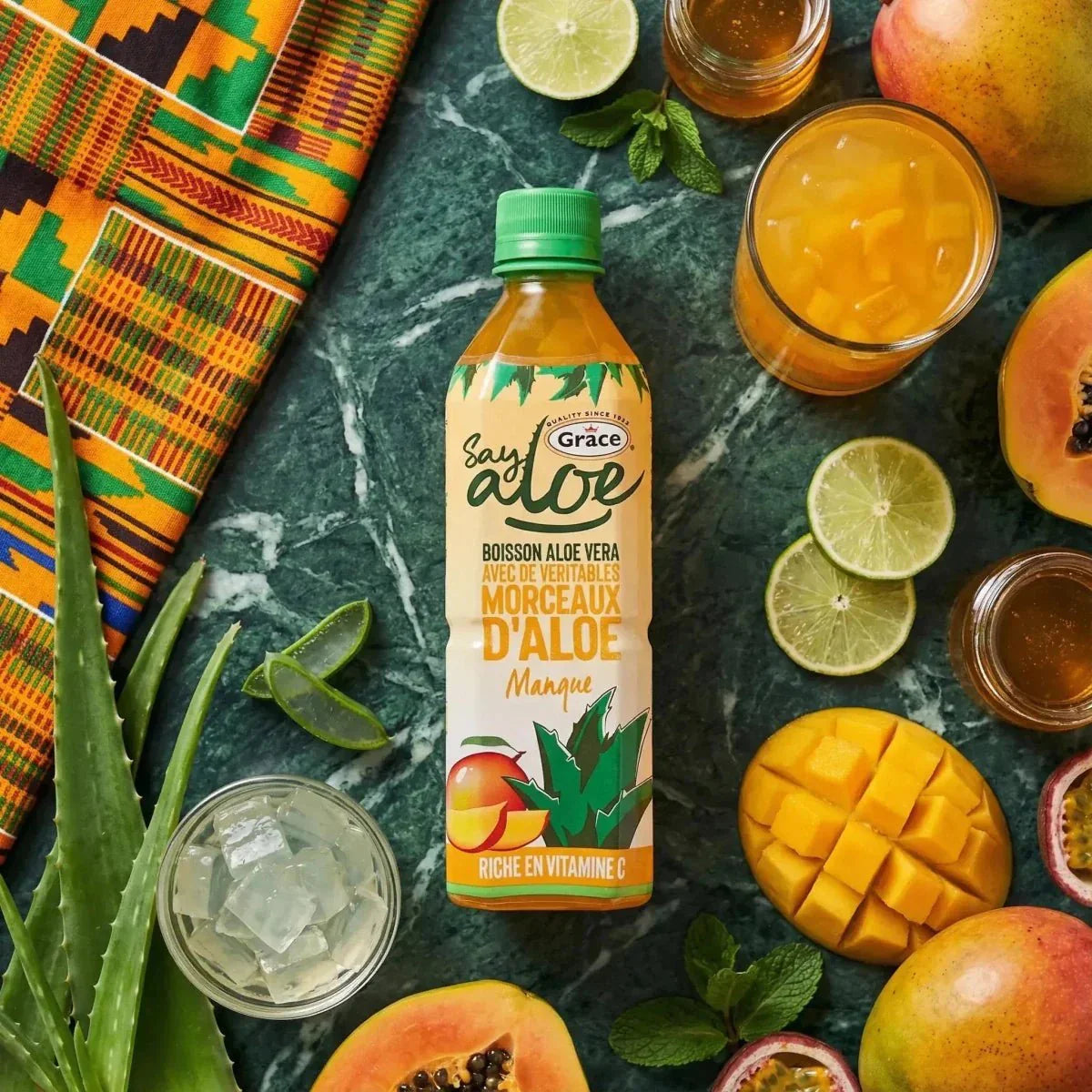 Close-up van een fles Grace Say Aloe mango omringd door tropisch fruit.