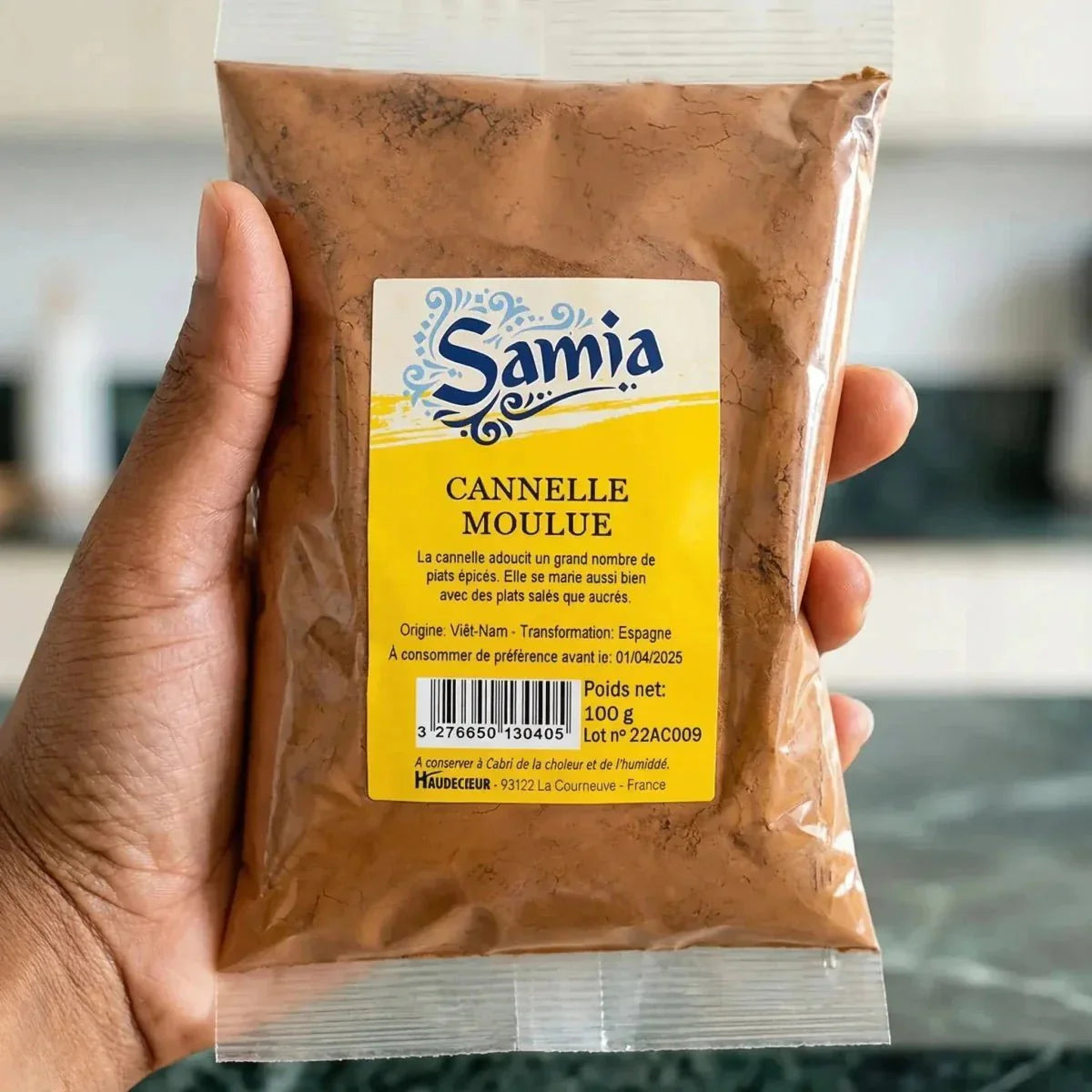 Un sachet de cannelle moulue Samia tenu par une main. L'emballage est jaune et transparent.