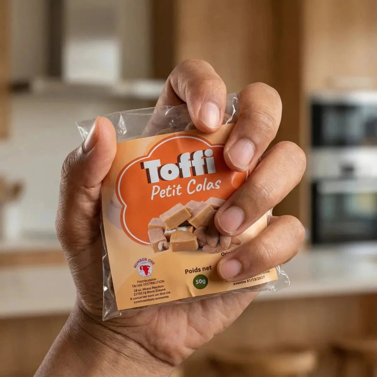 Gros plan d'une main tenant un sachet de caramels Toffi Petit Colas. Emballage orange.