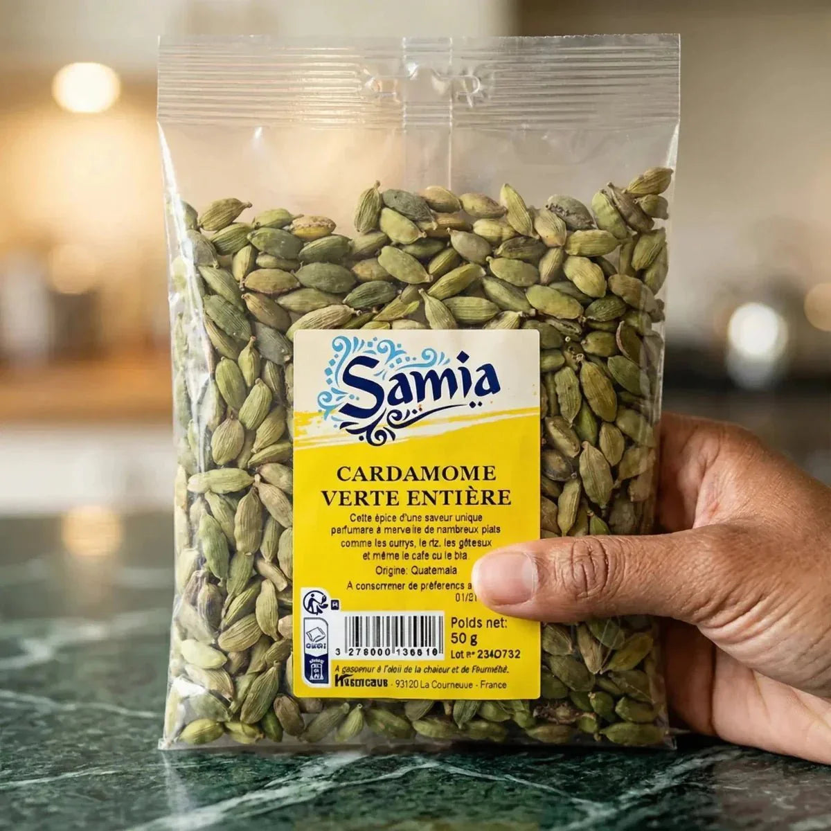 Sachet de cardamome verte entière SAMIA, 50g, tenu en main devant un plan de travail en marbre.