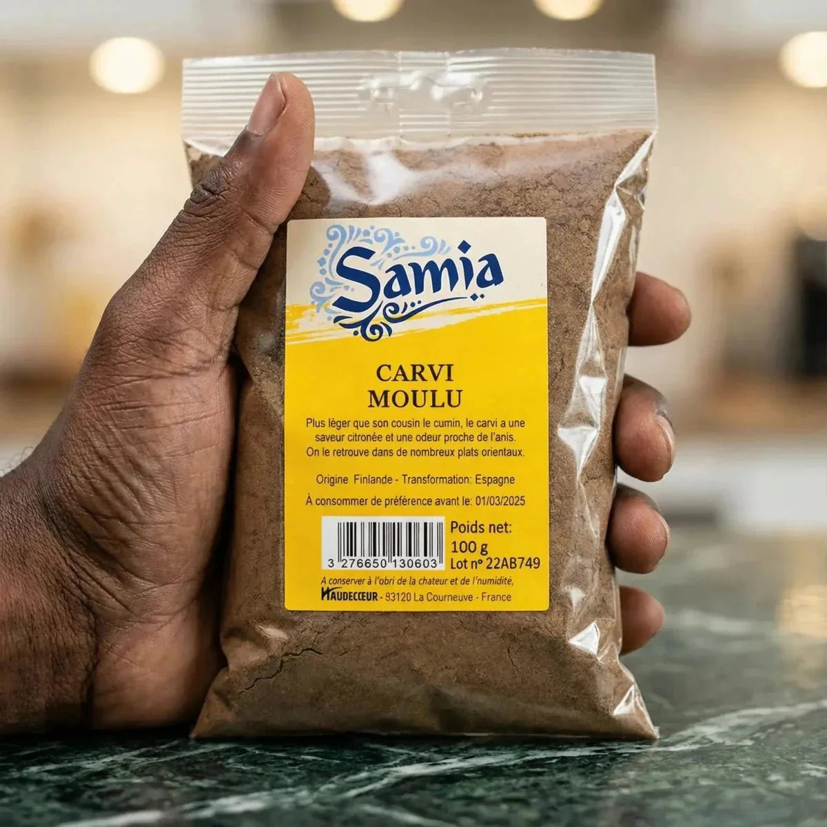 Un sachet de carvi moulu SAMIA, 100g, présenté à la main, sur un fond de marbre.