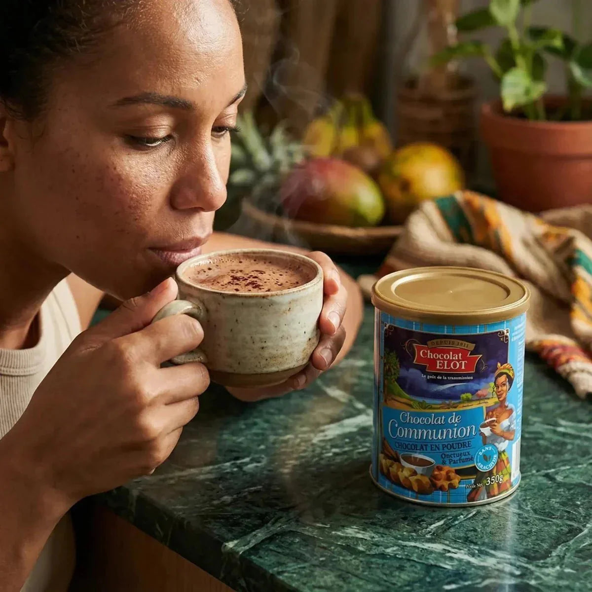 Vrouw die warme chocolademelk drinkt naast een doos Communion Chocoladepoeder CHOCOLAT ELOT.