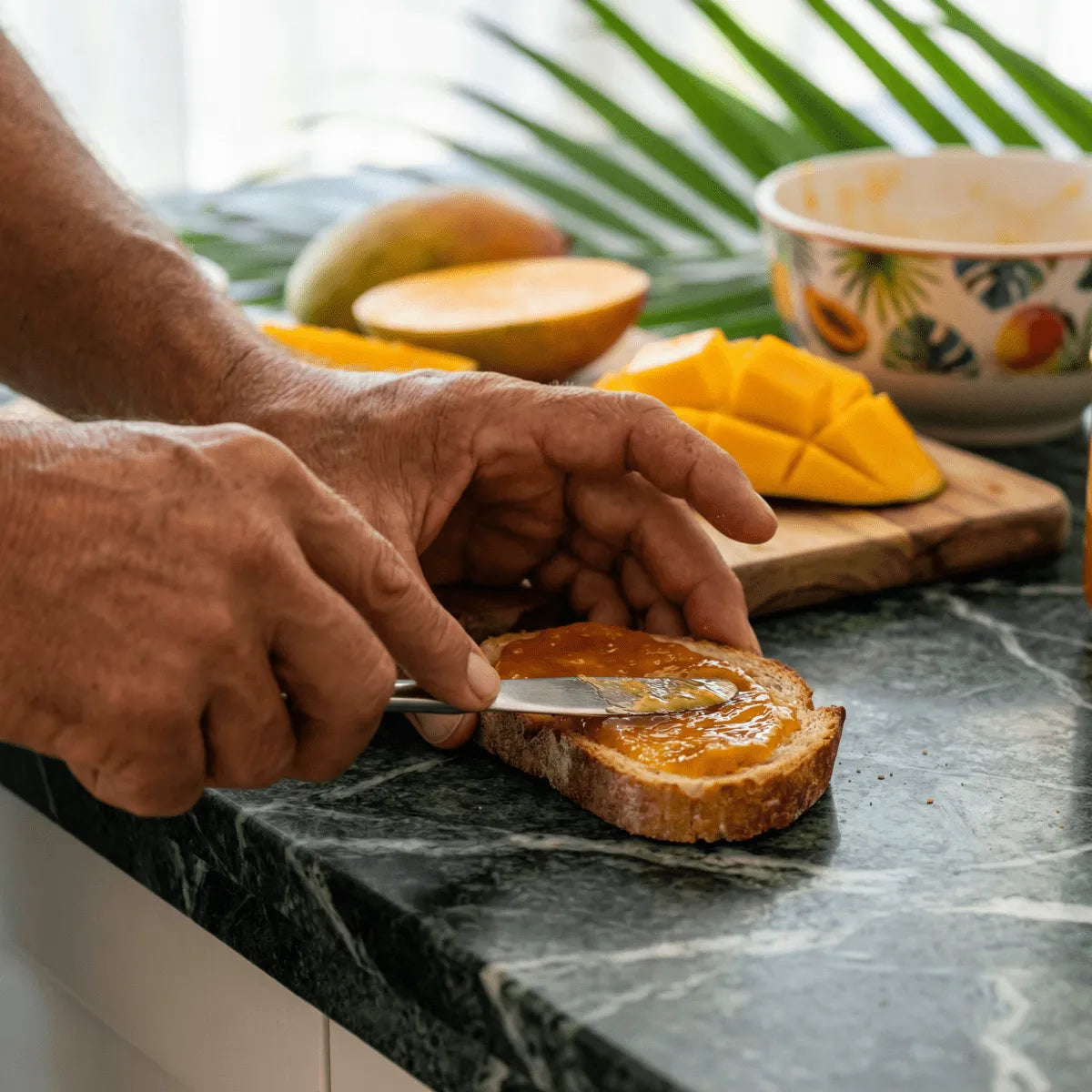 M'AMOUR mango jam on a slice of bread, fresh mangoes in the background.