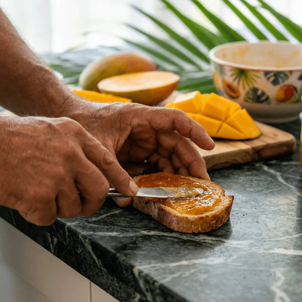 Confiture de mangue M'AMOUR sur une tartine, mangues fraîches en arrière-plan.