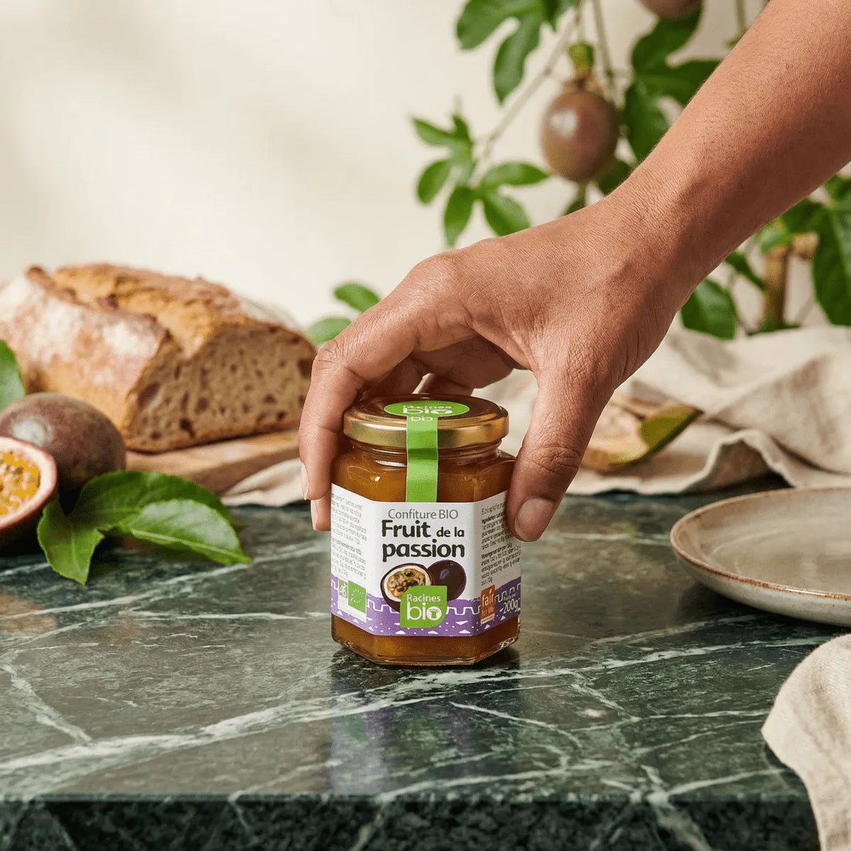 RACINES BIO passion fruit jam, 200g. A hand grabs the jar in front of some bread.