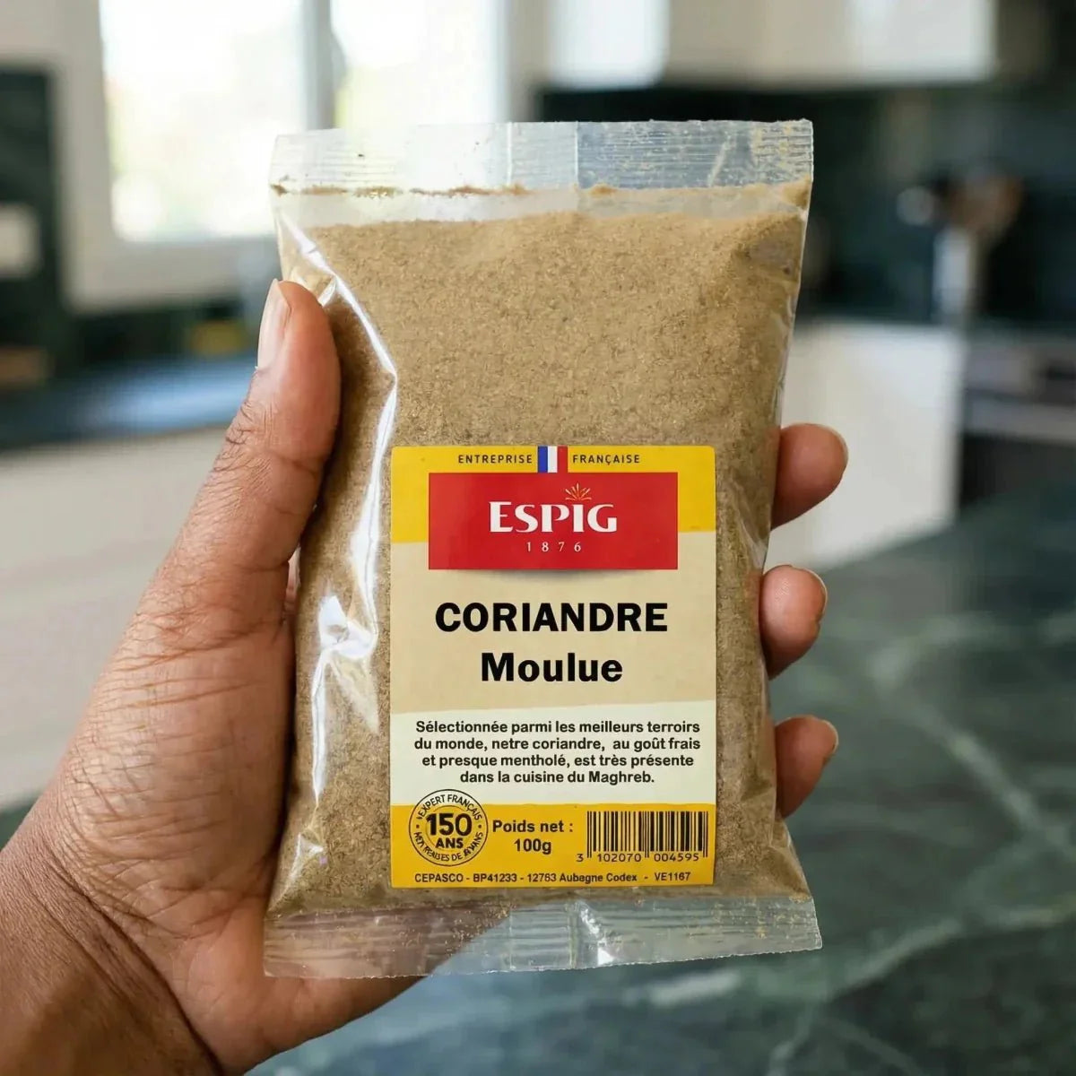 Handgehaltenes Beutelchen mit gemahlenem Koriander ESPIG (100g), Küche im Hintergrund.