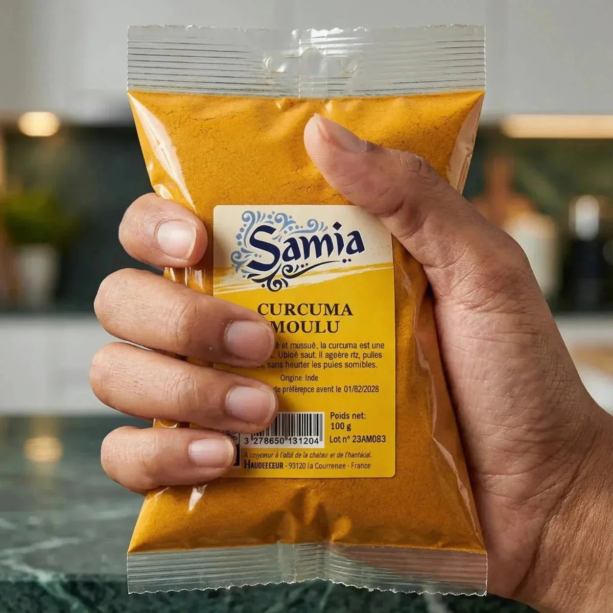 Bustina di curcuma macinata SAMIA tenuta in una mano su uno sfondo di cucina sfocato.