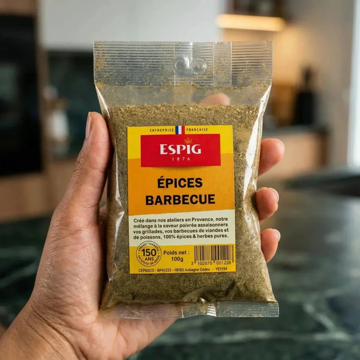 Un sachet d'épices barbecue ESPIG tenu à la main, avec une cuisine floue en arrière-plan.