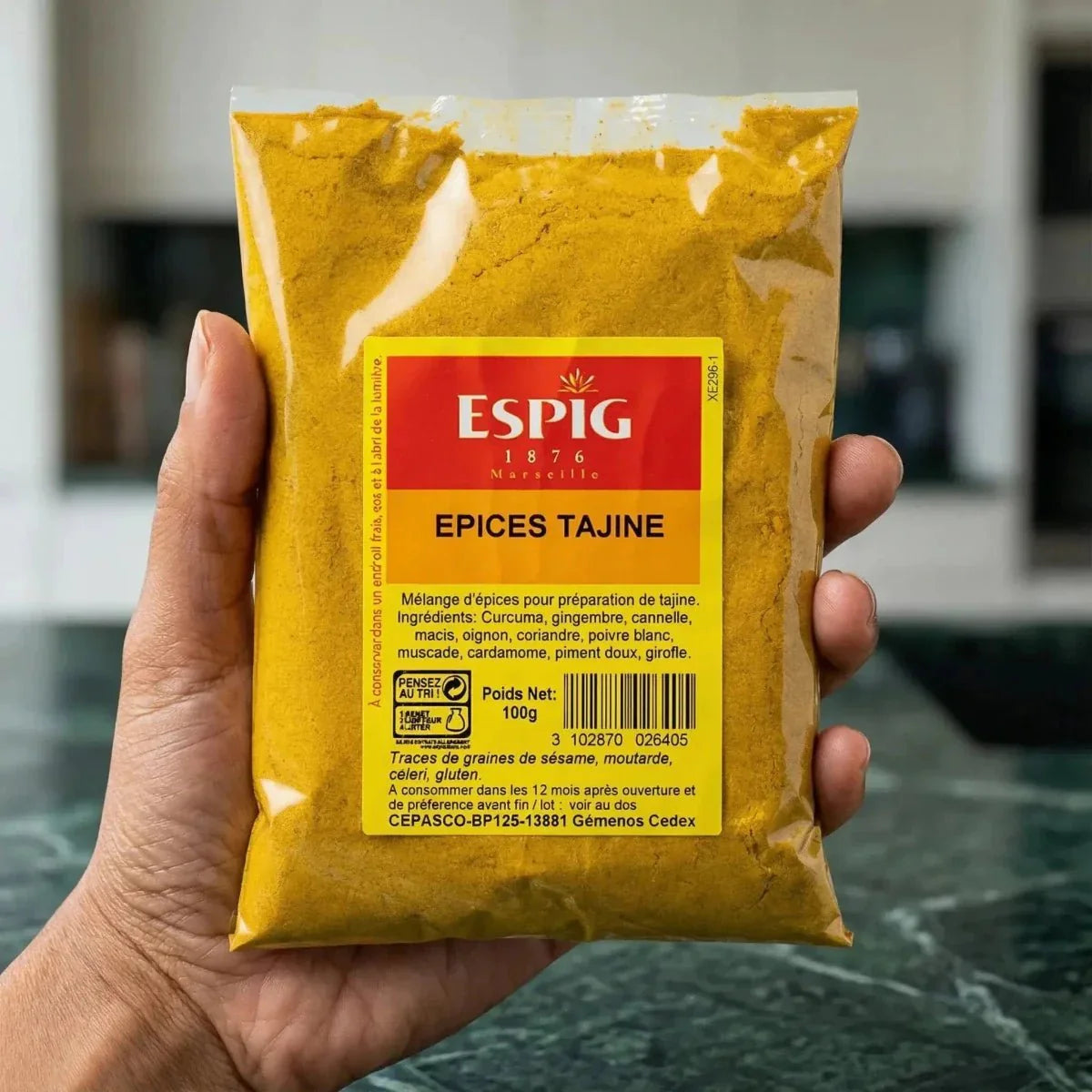 Hand holding a 100g ESPIG Tajine spice sachet. Yellow and red packaging, kitchen in the background.