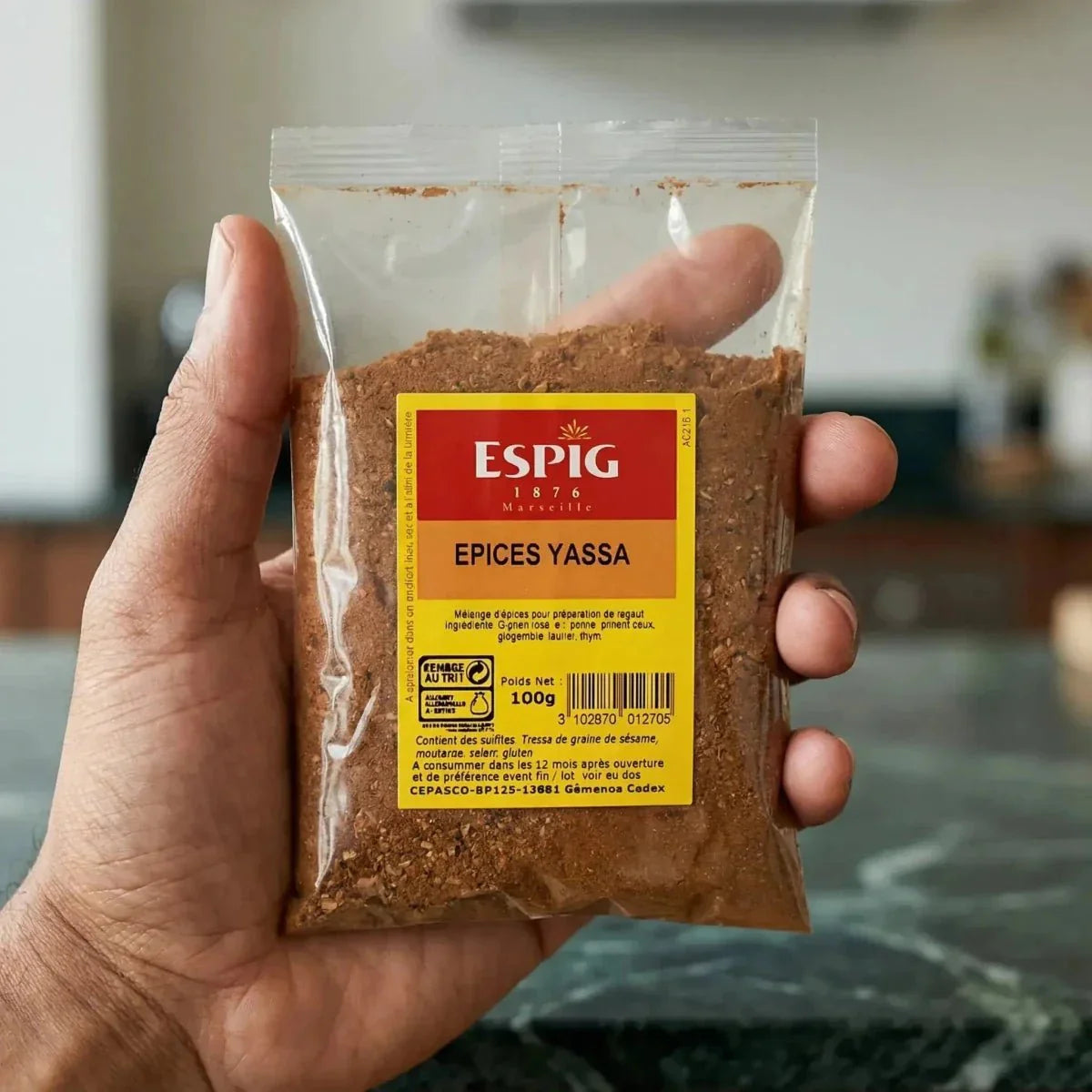 A 100g bag of ESPIG Yassa Spice held in a hand against a blurred kitchen background.