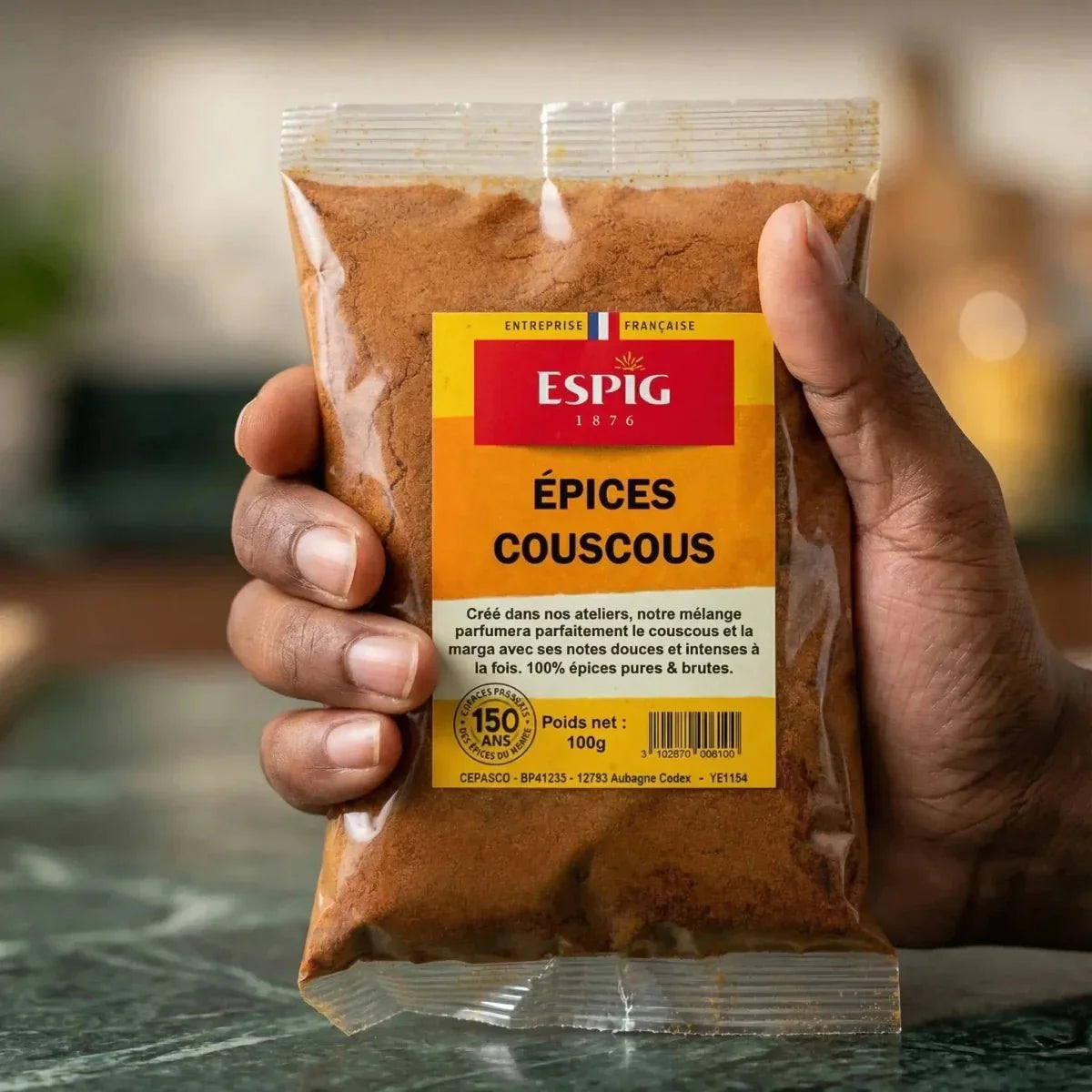Un sachet d'Épices Couscous ESPIG tenu en main, sur un fond de plan de travail vert marbré.
