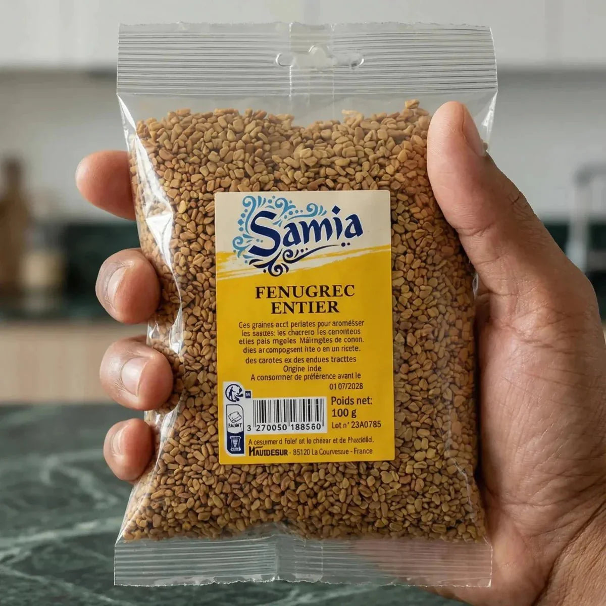 Paket mit ganzem Bockshornklee Samia 100g, in einer Hand gehalten vor einem Küchenhintergrund.
