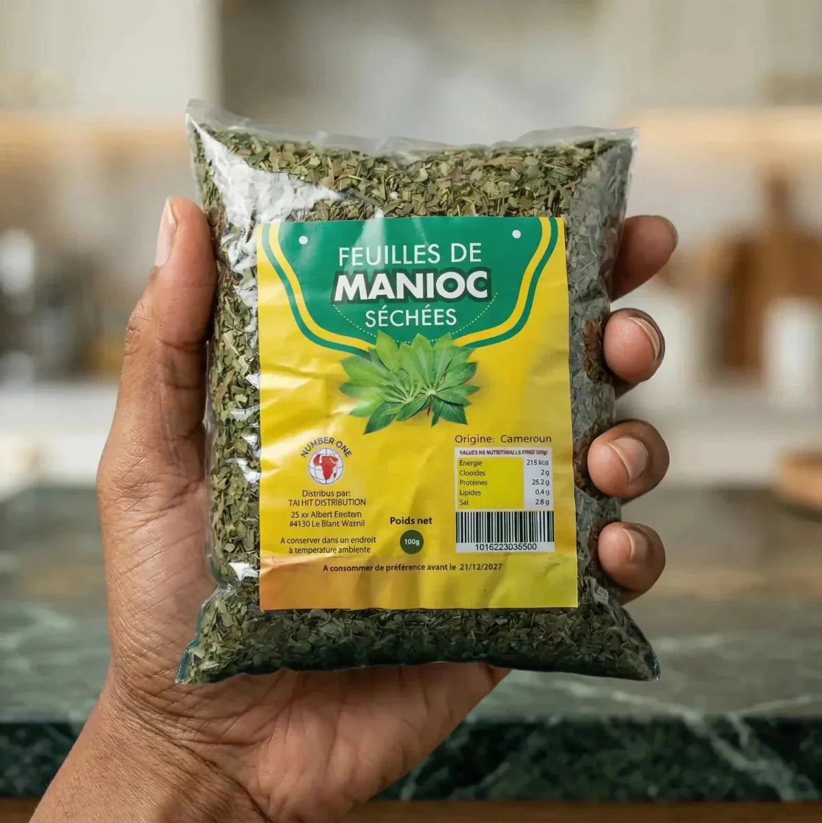 Dried cassava leaves NUMBER ONE, 100g, held by a hand. Yellow packaging.