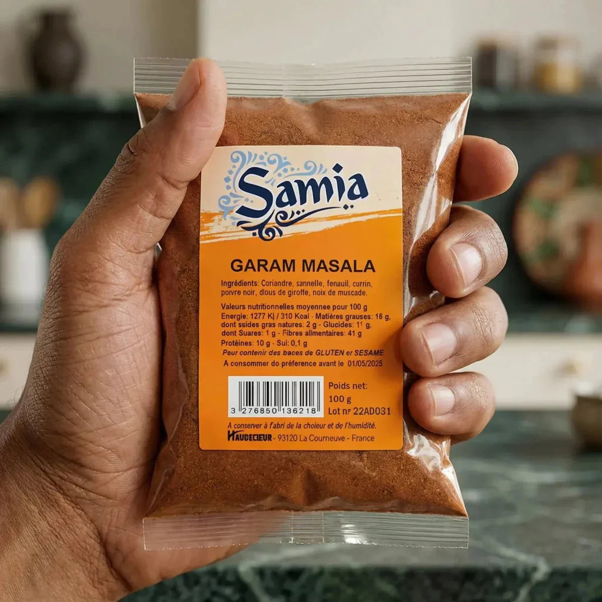 Una bustina di garam masala Samia da 100g mostrata in una mano, davanti a uno sfondo di cucina.