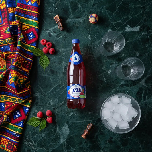 Bouteille de Soda Framboise GAZOUZ posée sur une table avec glaçons, verres et framboises, boisson pétillante fruitée et désaltérante.
