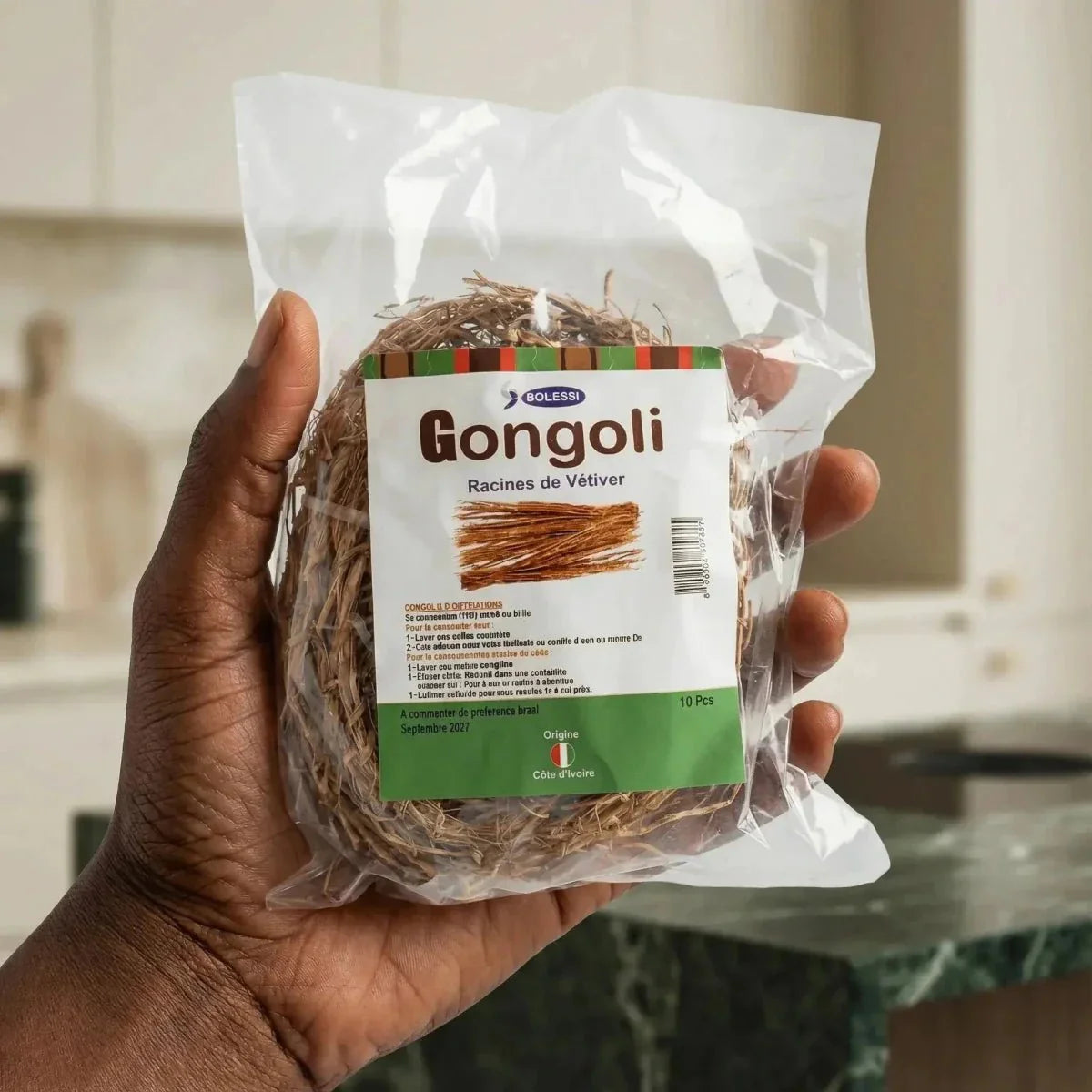 Gongoli Bolessi, racines de vétiver en sachet tenu dans une main. Étiquette bilingue.