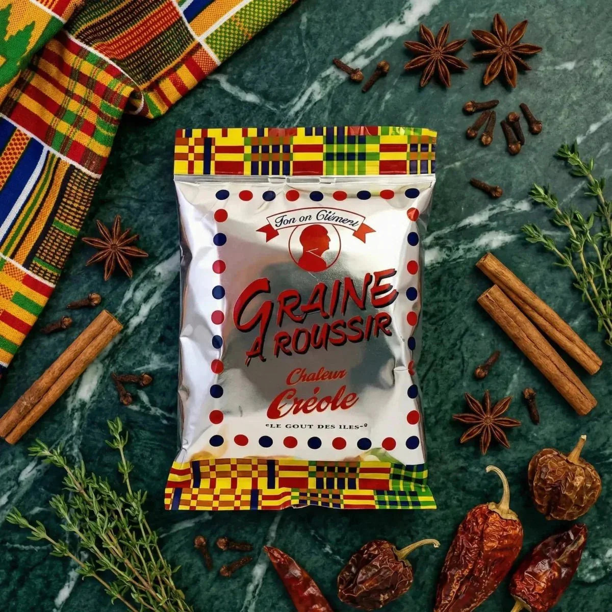Tonton Clément roasting seed surrounded by spices and a colorful fabric on a marbled green background.