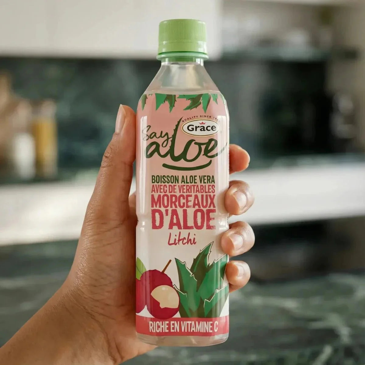 Een hand houdt een fles Aloë- en Litchisap van GRACE vast. Keukenachtergrond.