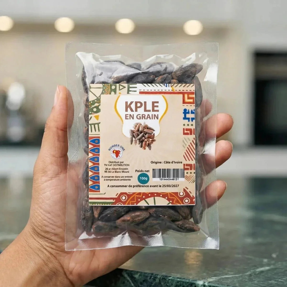 Hand holding a 100g sachet of whole NUMBER ONE Kpele, packaging with African patterns.