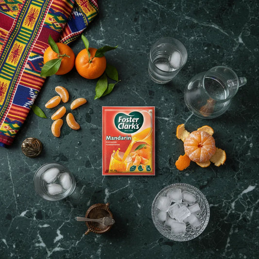 Sachet de jus mandarine FOSTER CLARKS entouré de verres, glaçons et mandarines fraîches sur une table, boisson fruitée idéale pour l'été.