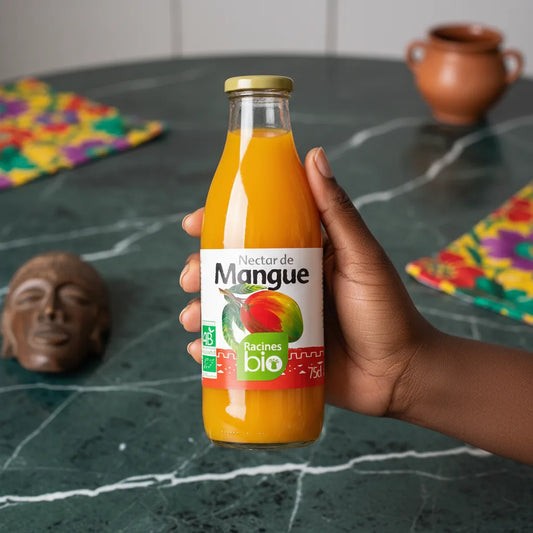 Main tenant une bouteille de Nectar de Mangue Bio Racines, boisson bio sucrée et veloutée, sur une table verte avec décor africain.