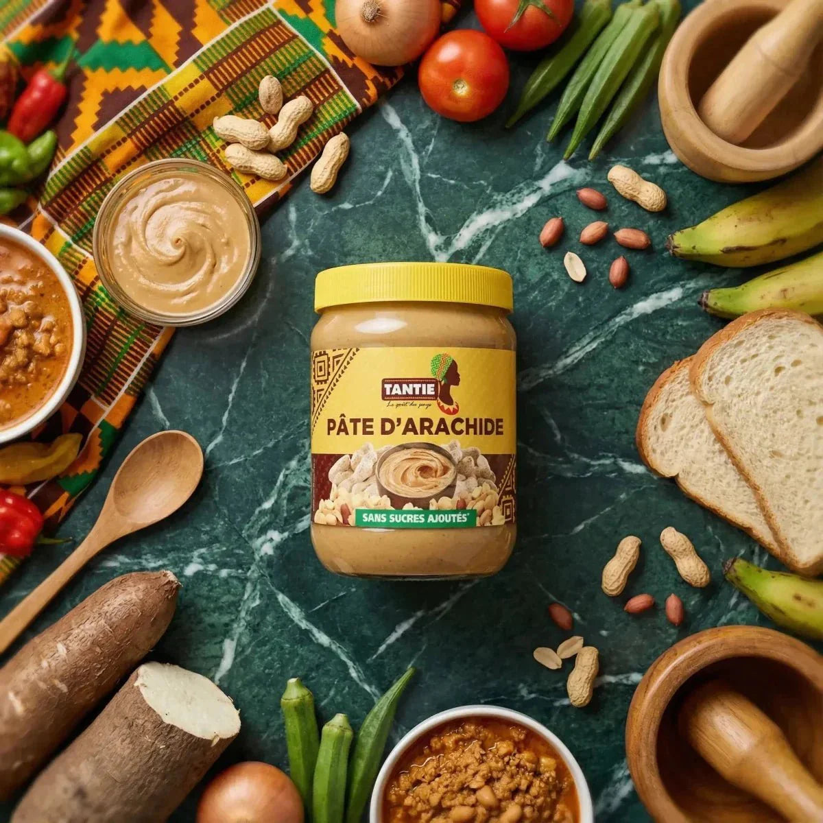 Erdnussbutter TANTIE 500g auf einem Tisch mit Erdnüssen, Gemüse, Brot und Mörsern.