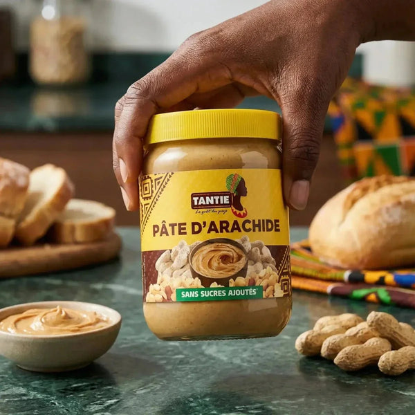 Pâte d'arachide crémeuse par TANTIE, avec des arachides entières, pot jaune tenu dans la main.
