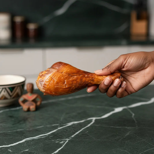 Main tenant un pilon de dinde fumée sur plan de travail, illustrant la texture riche et le fumage artisanal de cette volaille surgelée.