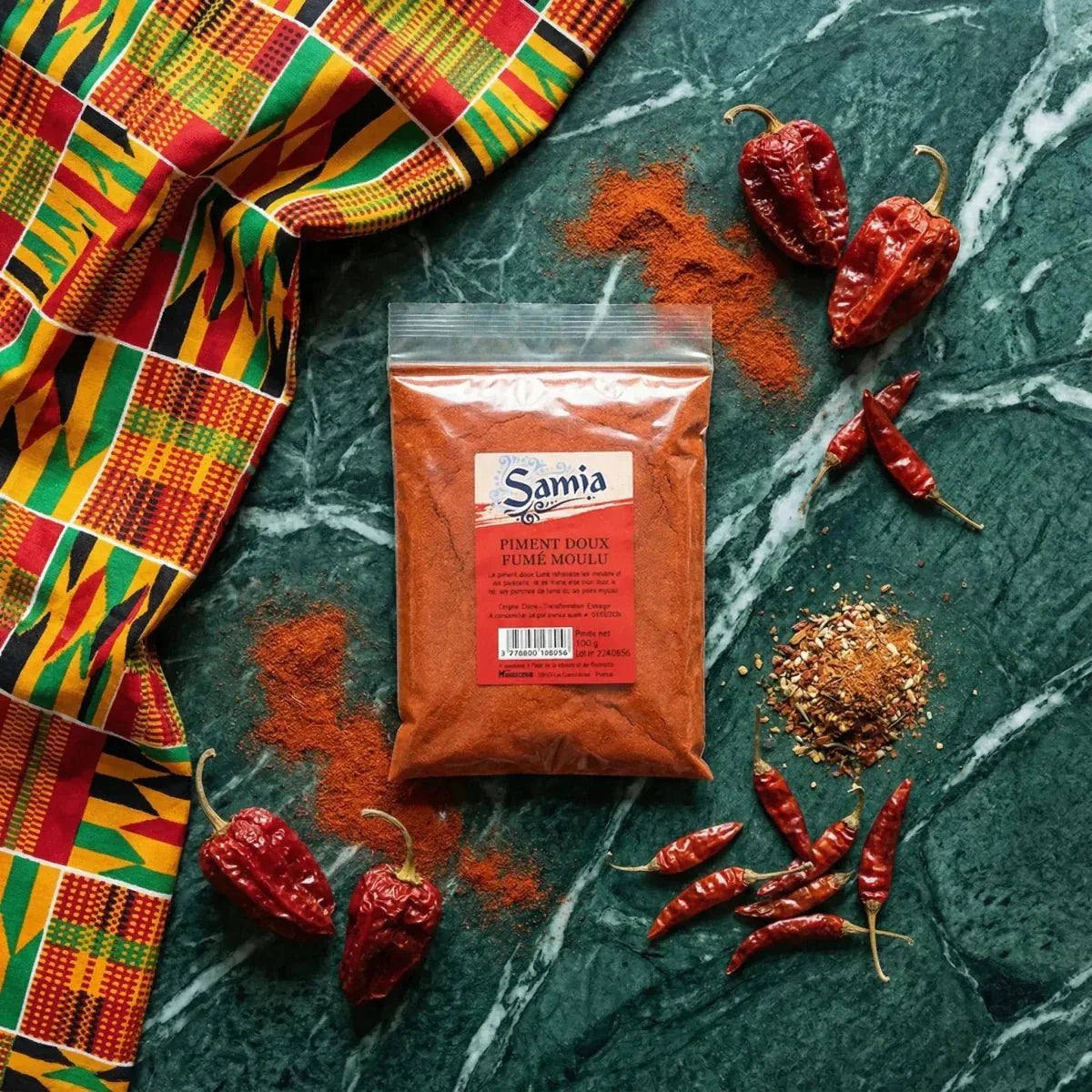 Bustina SAMIA di peperoncino dolce affumicato macinato, circondata da peperoncini rossi interi e polvere marmorizzata.