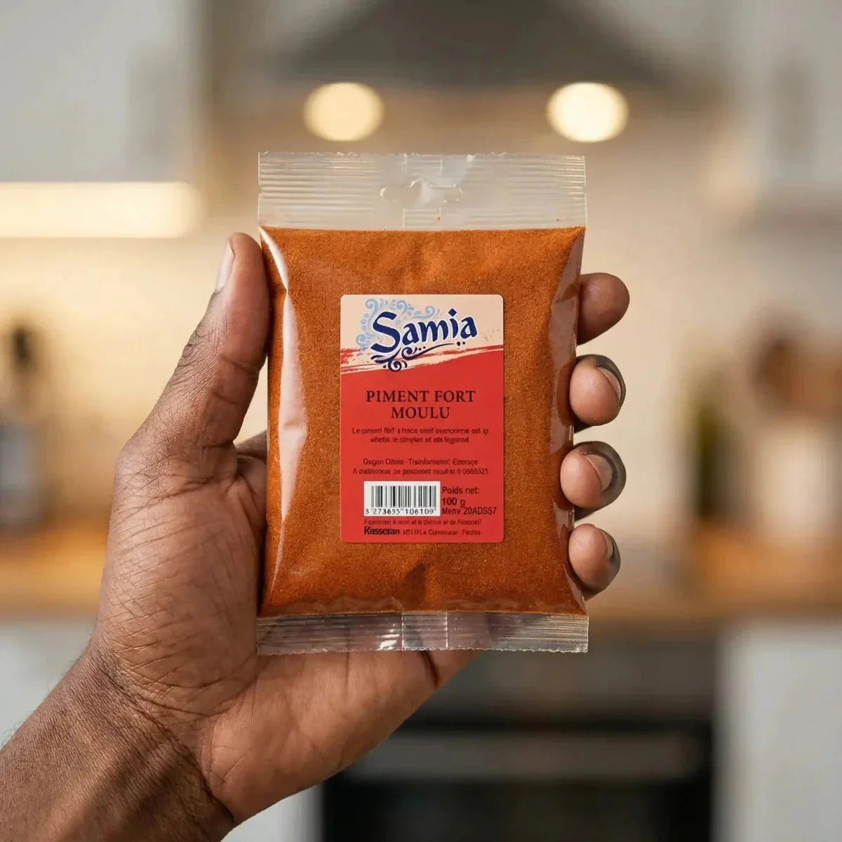 Close-up of a hand holding a packet of SAMIA ground hot chili. Blurred kitchen background.
